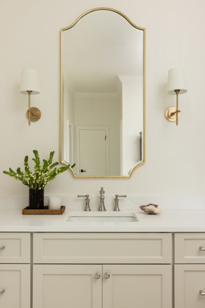 upscale neutral bathroom featuring a brass mirror, brass sconces on either side of the mirror, and polished nickel faucet and marble counter